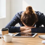 Mujer apoya la cabeza sobre su escritorio, agotada, junto a un computador y una taza de café